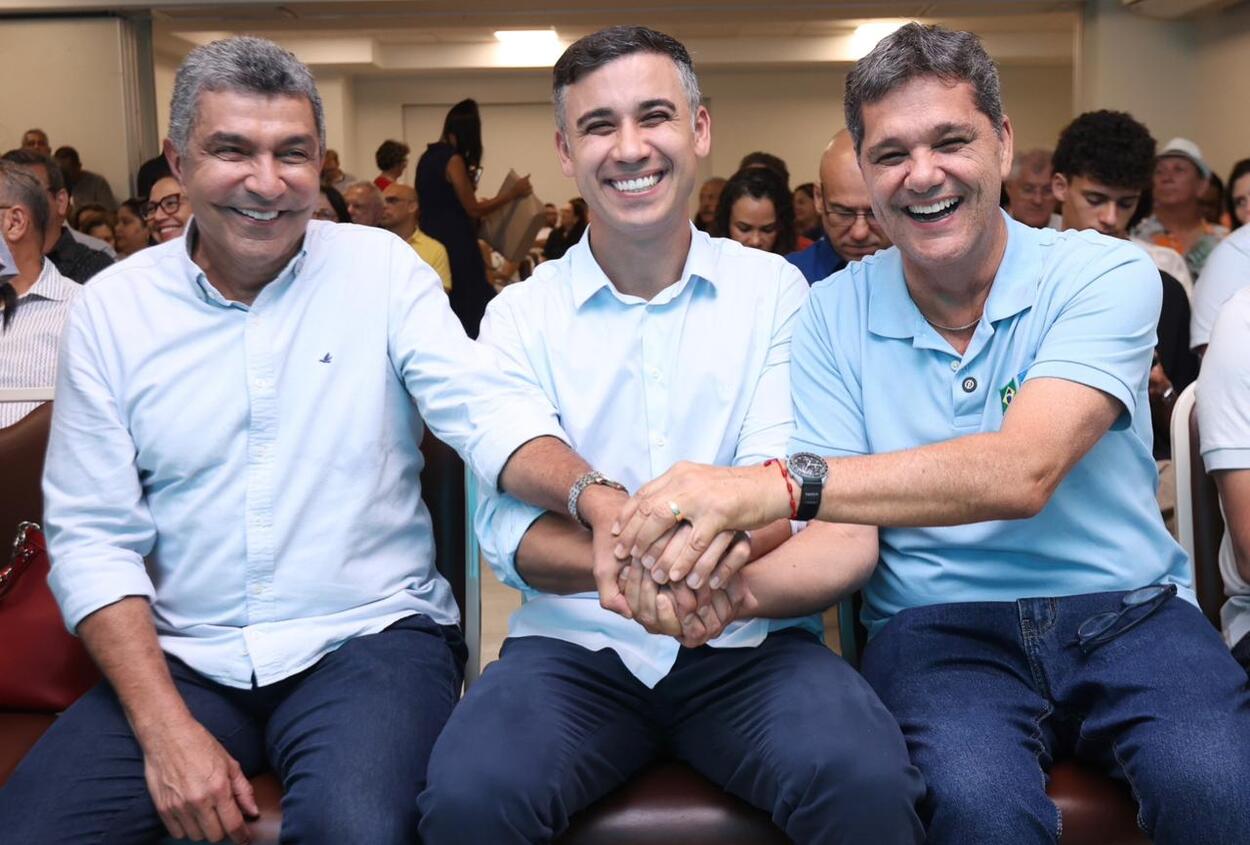 Três homens sentados lado a lado em um auditório lotado sorriem enquanto apertam as mãos em gesto de união. Eles vestem camisas claras e parecem participar de um evento público, com várias pessoas ao fundo observando.