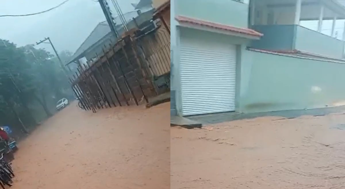 Rua tomada por enchente em dia de chuva forte. A água barrenta cobre completamente a via, que aparece inclinada na foto. À esquerda, algumas motos estão paradas; à direita, há casas e muros. No fundo, um carro branco está parcialmente submerso. A chuva intensa reduz a visibilidade e reforça o cenário de alagamento.