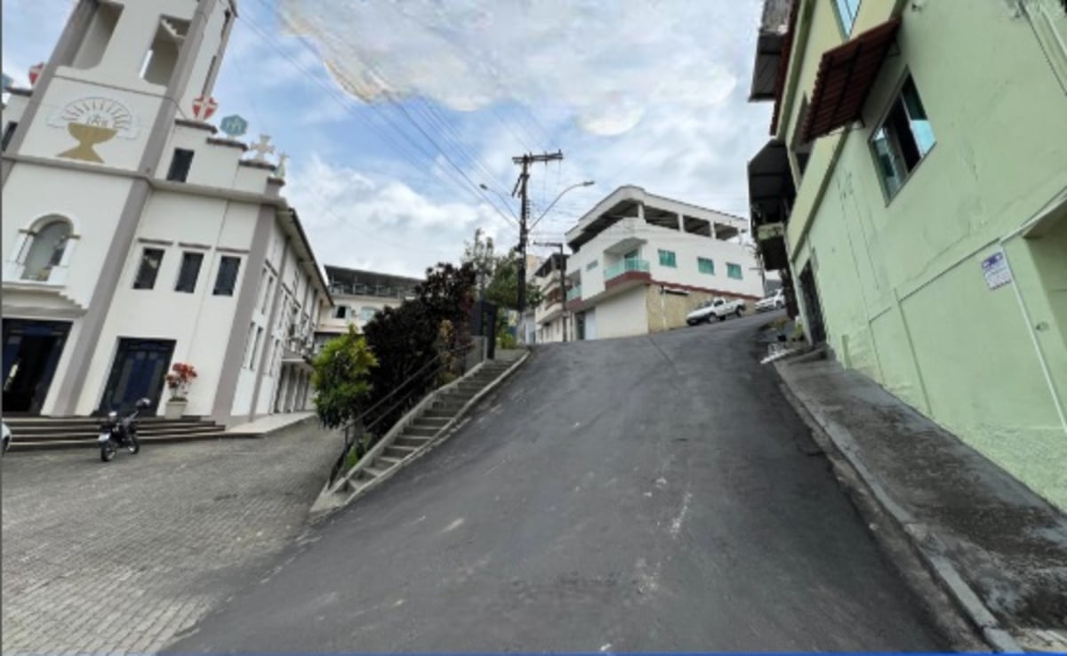 Rua íngreme em área urbana, com calçada lateral e escadaria ao centro. À esquerda, há um prédio religioso de fachada branca com detalhes em bege. À direita, um prédio verde claro com janelas. No topo da ladeira, aparecem casas e um carro estacionado. O céu está nublado.