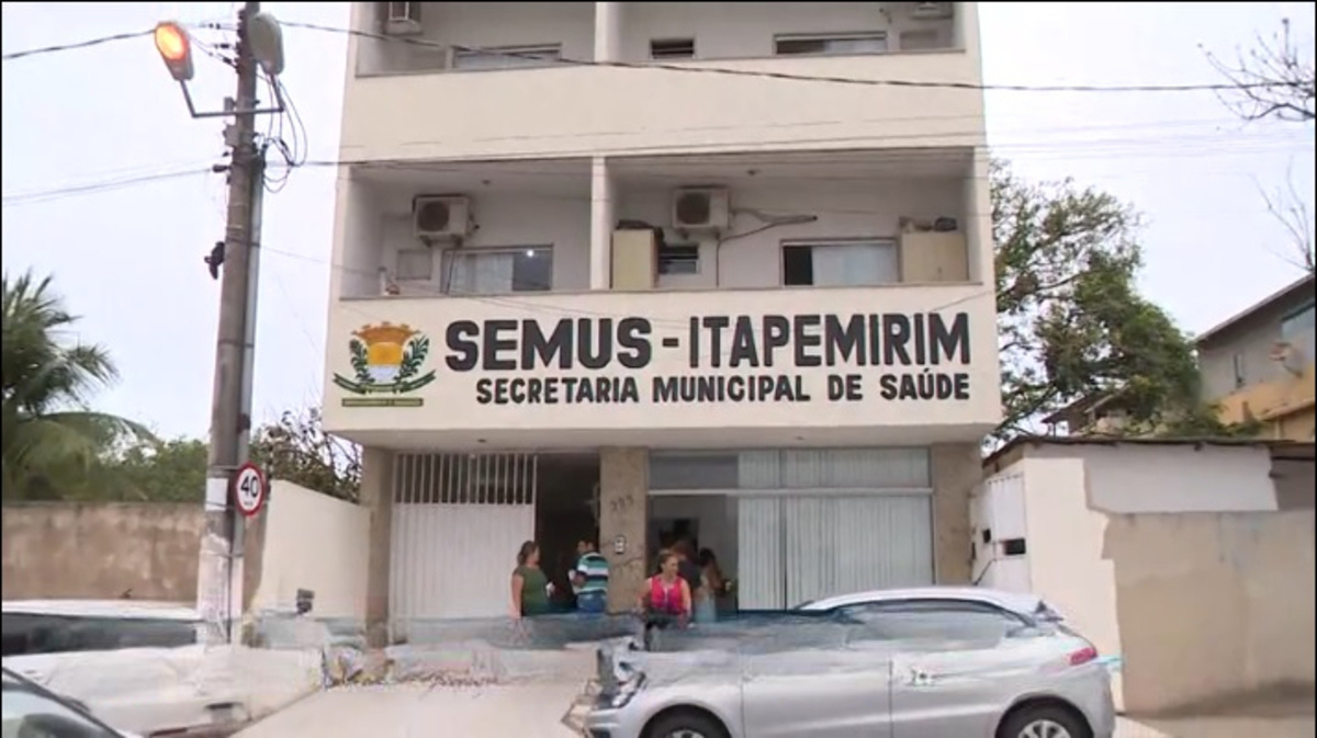 Fachada da Secretaria Municipal de Saúde de Itapemirim (SEMUS-Itapemirim). O prédio é de três andares, com letreiro grande na parte frontal indicando o nome da instituição. Em frente à entrada, algumas pessoas estão reunidas na calçada. Há carros estacionados na via e um poste com placa de limite de velocidade de 40 km/h ao lado esquerdo da imagem.