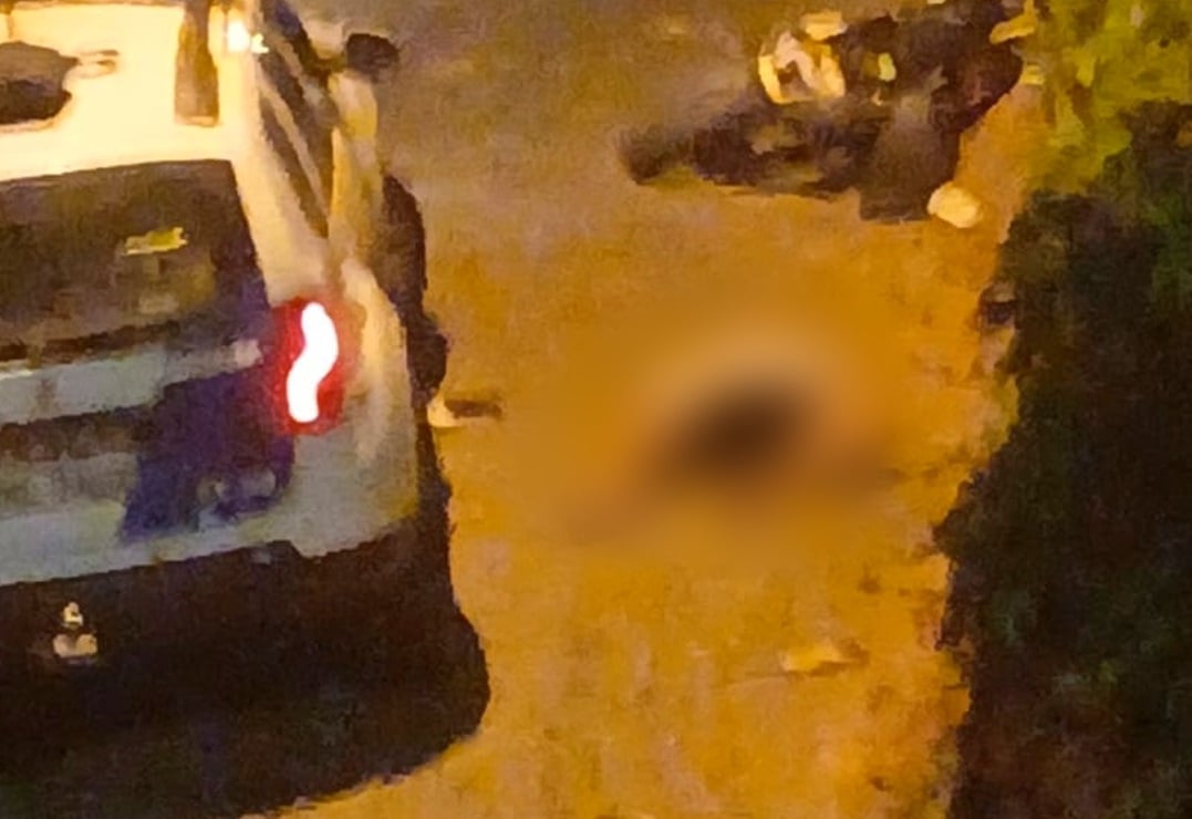 Viatura policial parada em rua de paralelepípedos durante a noite, com uma motocicleta caída ao fundo. Uma área desfocada ao centro indica o local onde um suspeito foi encontrado após intervenção policial. A iluminação amarelada das lâmpadas de rua destaca o cenário cercado por árvores e sombras.