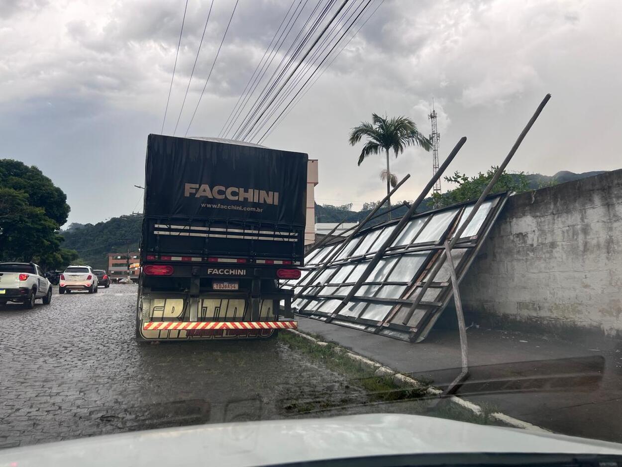 A imagem mostra uma rua de paralelepípedos molhada após a chuva, com um caminhão da marca Facchini parado ou trafegando lentamente no lado esquerdo. À direita, uma grande estrutura metálica, semelhante a um painel ou cerca, está caída e apoiada sobre um muro, aparentando ter sido derrubada pelo vento. Há carros seguindo pela via, postes e fios de energia ao longo da rua, além de palmeiras e morros ao fundo sob um céu nublado e carregado, indicando tempo instável.