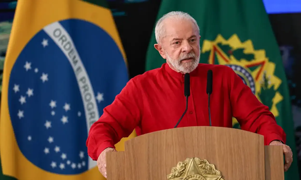 Homem mais velho, de barba grisalha e camisa vermelha, fala em um púlpito com brasão dourado, diante de microfones. Ao fundo, há bandeiras do Brasil e outra bandeira verde com símbolo oficial.