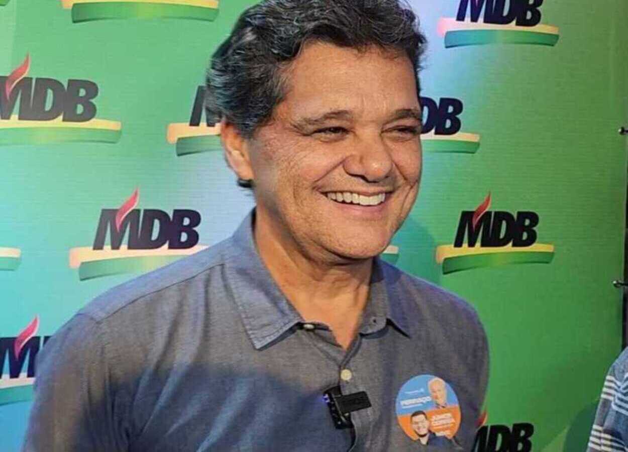 Vice-governador Ricardo Ferraço sorri durante evento político, em frente a painel verde com logotipo do MDB, usando camisa azul e adesivo de campanha no peito.