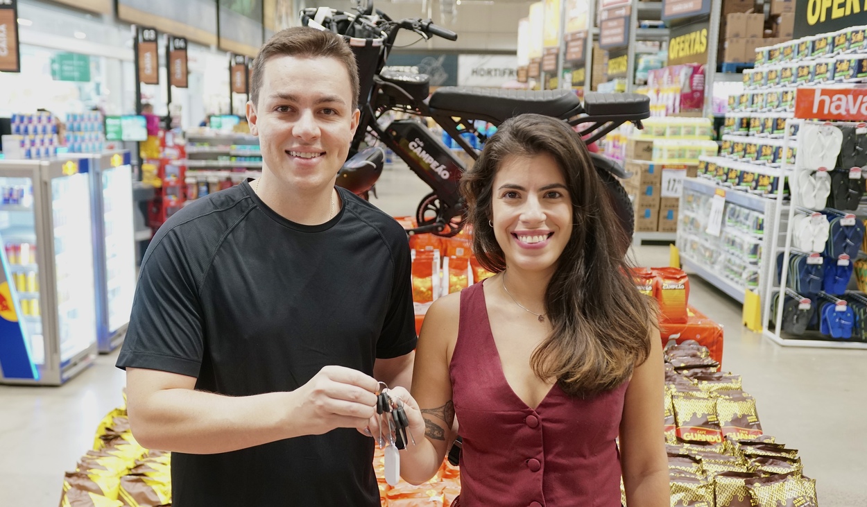 Cliente recebe chave simbólica durante entrega de prêmio em supermercado, ao lado de uma bicicleta elétrica exposta no local.