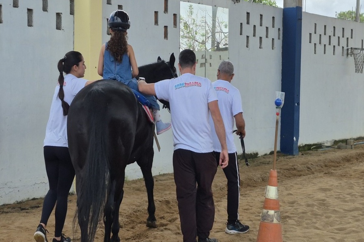 Criança com capacete participa de sessão de equoterapia montada em um cavalo preto, acompanhada por três profissionais em área de areia.