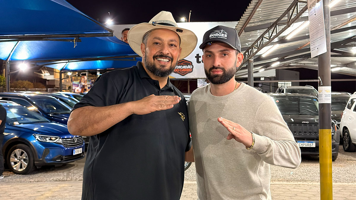 Fotografia noturna de dois homens posando lado a lado em um pátio iluminado de venda de carros. À esquerda está o Pastor da Uai, sorridente, usando um chapéu claro estilo boiadeiro e camisa polo preta. À direita está Alexandre, vestindo um boné preto com a logo "Borges Veículos e Caminhões" e uma blusa cinza de manga longa. Ambos fazem um gesto característico com as mãos espalmadas na altura do peito. Ao fundo, veem-se vários carros estacionados sob uma estrutura metálica com toldos azuis e iluminação artificial.