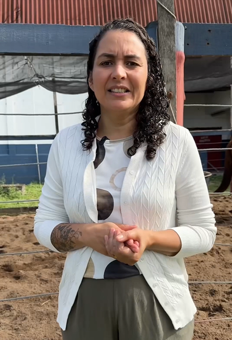 Mulher em pé, usando casaco branco, fala em área de equoterapia com arena de areia e estrutura coberta ao fundo.