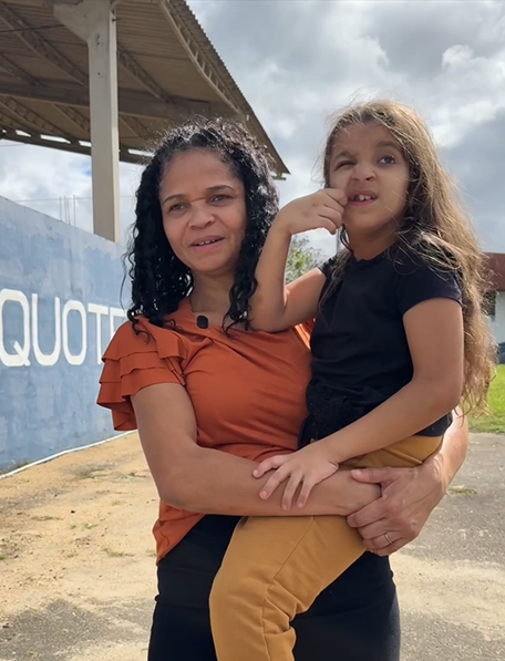 Mulher segura uma criança no colo em área externa de equoterapia, com muro azul e estrutura coberta ao fundo.