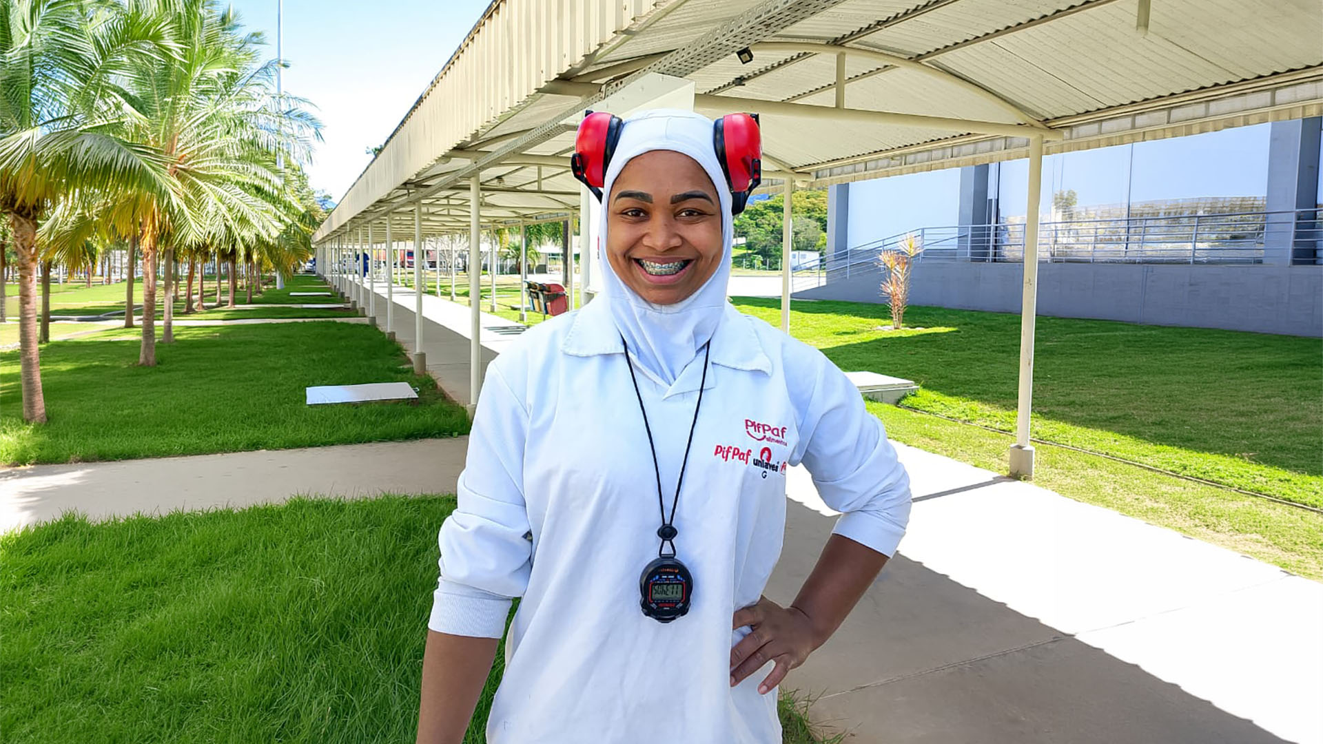 Mulher sorridente, usando uniforme branco, touca e protetores auriculares vermelhos, posa em área externa de uma unidade industrial, cercada por gramado e palmeiras.