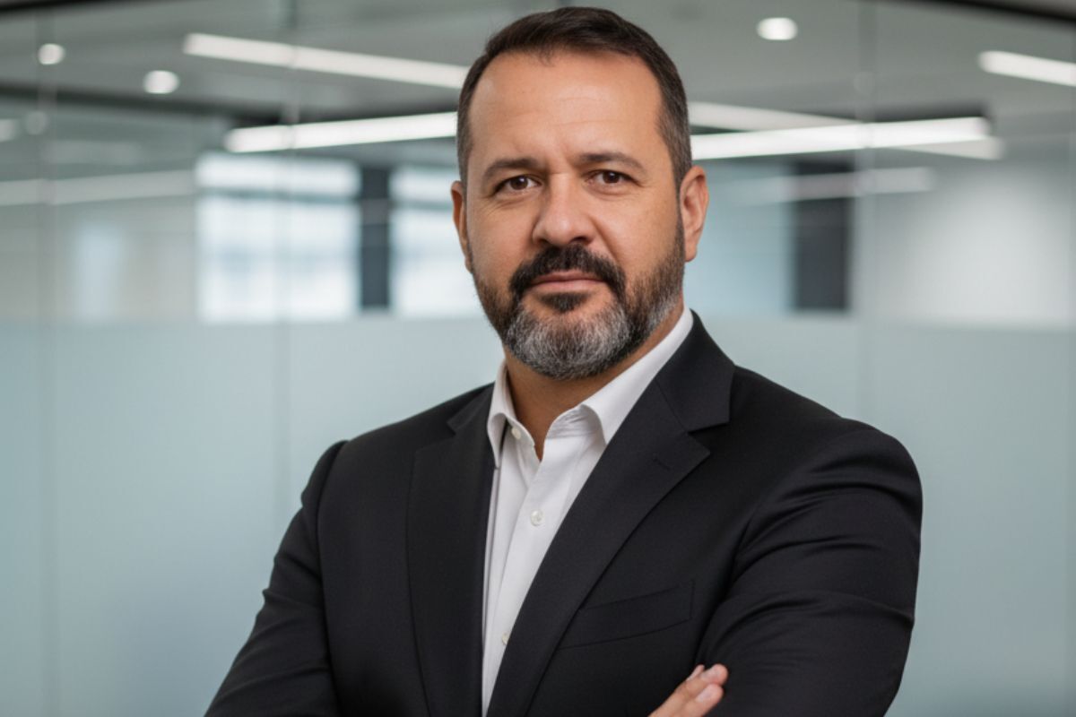 Homem de terno preto e camisa branca posa de braços cruzados em um escritório moderno.