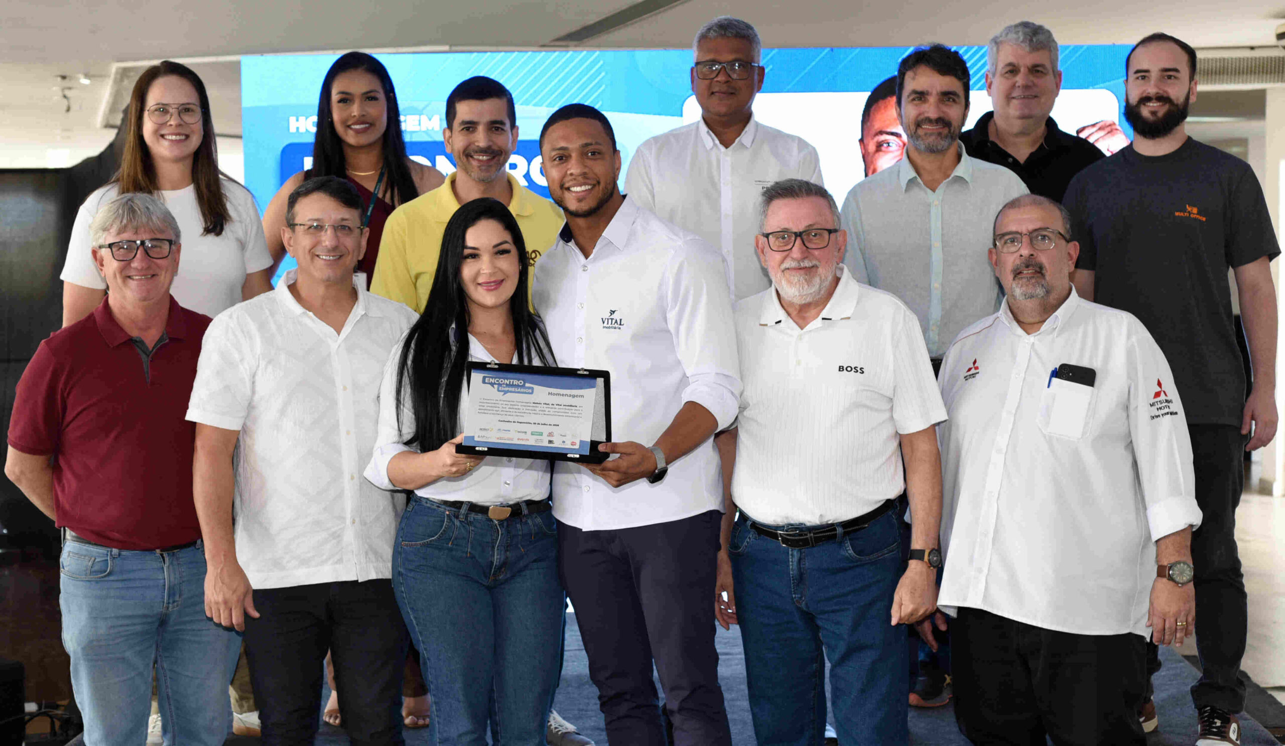 Grupo de empresários e convidados posa para foto durante evento, enquanto duas pessoas seguram uma placa de homenagem.