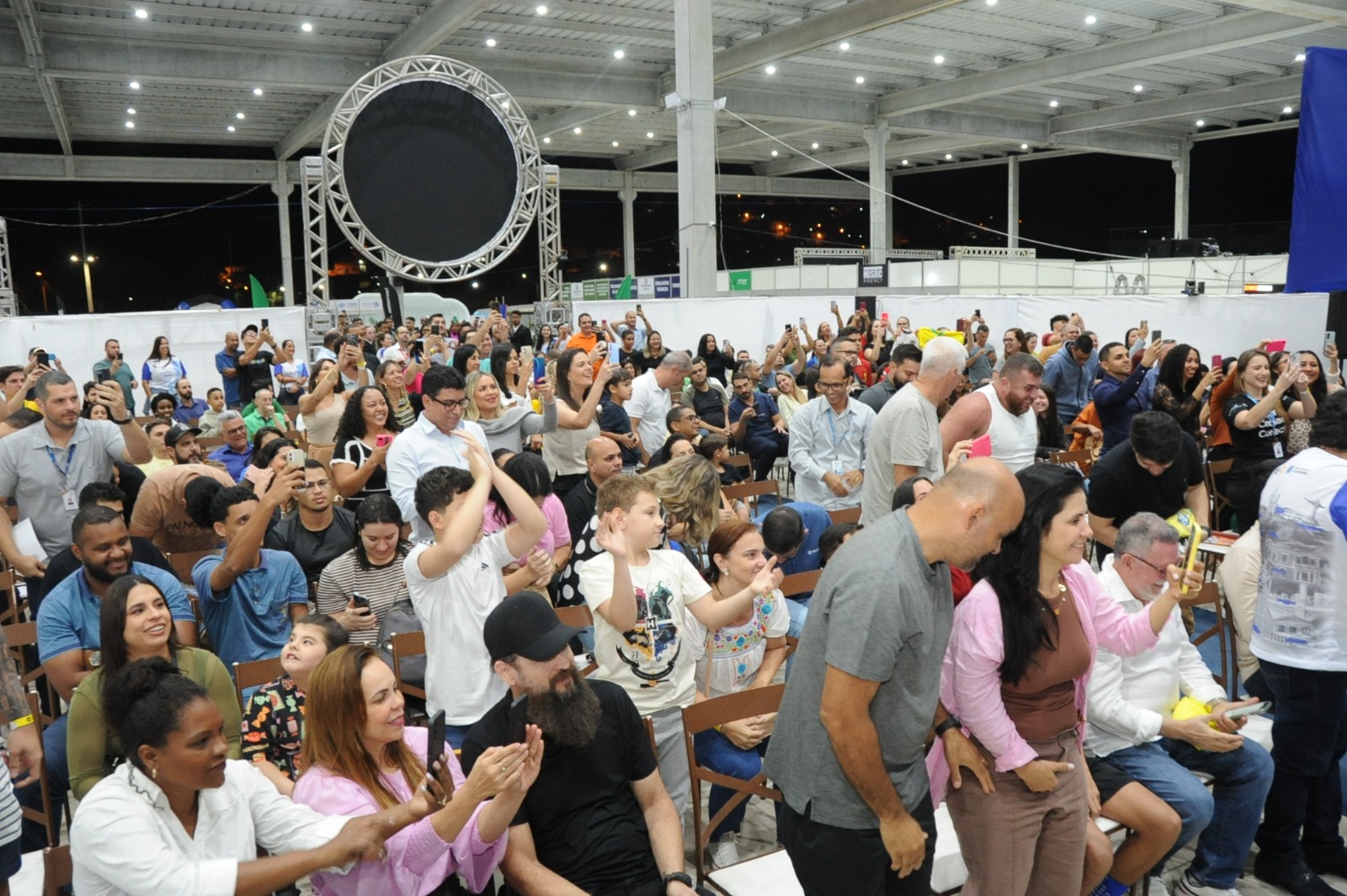 Público reunido em um pavilhão durante um evento, com pessoas em pé, aplaudindo e registrando o momento com celulares.