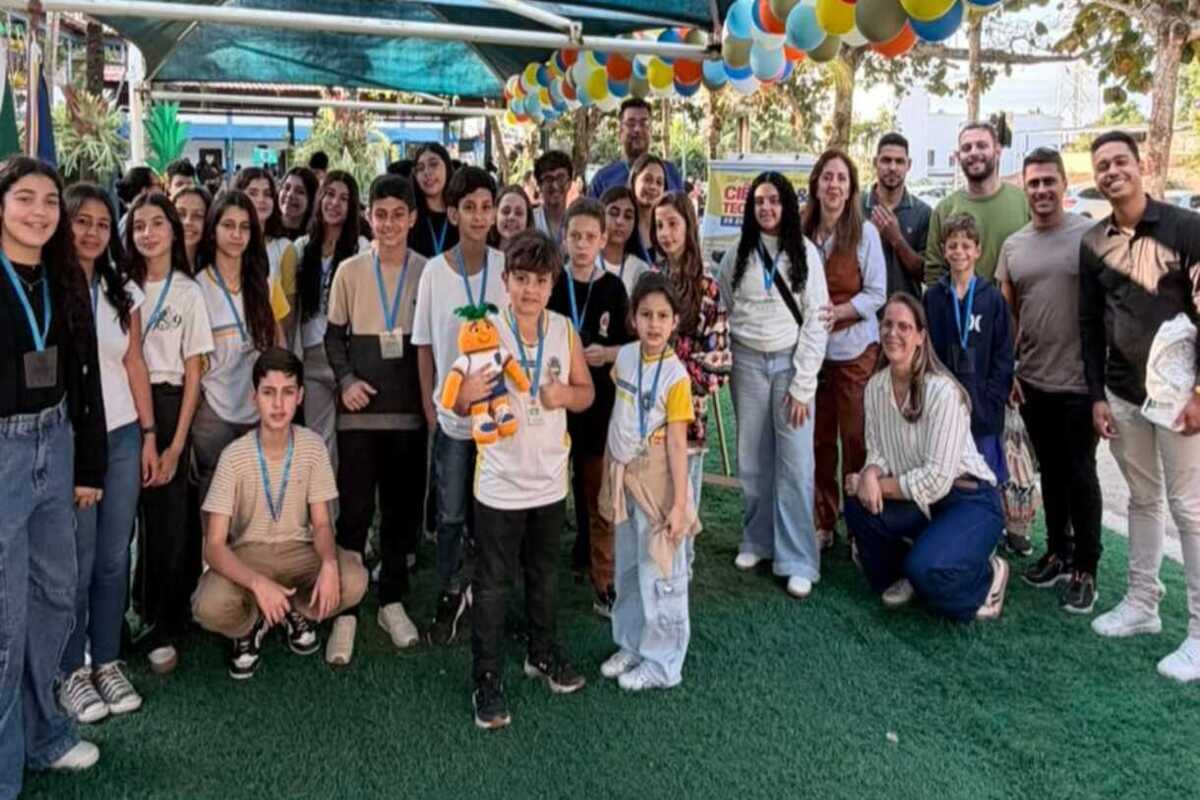 Grupo de estudantes e educadores posa para foto em uma feira de ciências, sob uma decoração com balões coloridos. Muitos participantes usam crachás e estão reunidos em uma área externa.