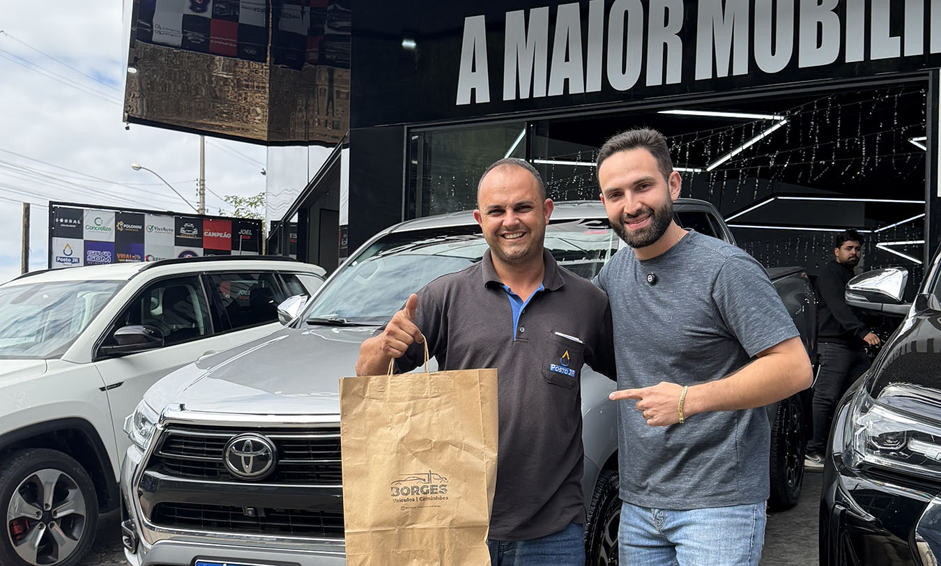 Dois homens sorriem para a câmara num stand de automóveis. O homem da esquerda veste um pólo cinzento, faz um sinal de polegar para cima com uma mão e com a outra segura um saco de papel com o logótipo da 'Borges Veículos e Caminhões'. O homem da direita veste uma t-shirt cinzenta e aponta sorridente para o saco. Atrás deles, está estacionada uma carrinha pick-up Toyota prateada e vê-se a fachada preta do estabelecimento com o letreiro parcial 'A MAIOR MOBILI'.