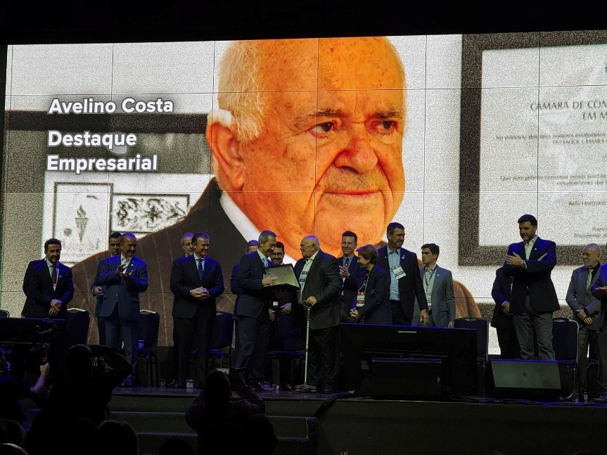Grupo de pessoas participa de uma homenagem no palco, diante de um telão com a imagem de Avelino Costa e a frase “Destaque Empresarial”.